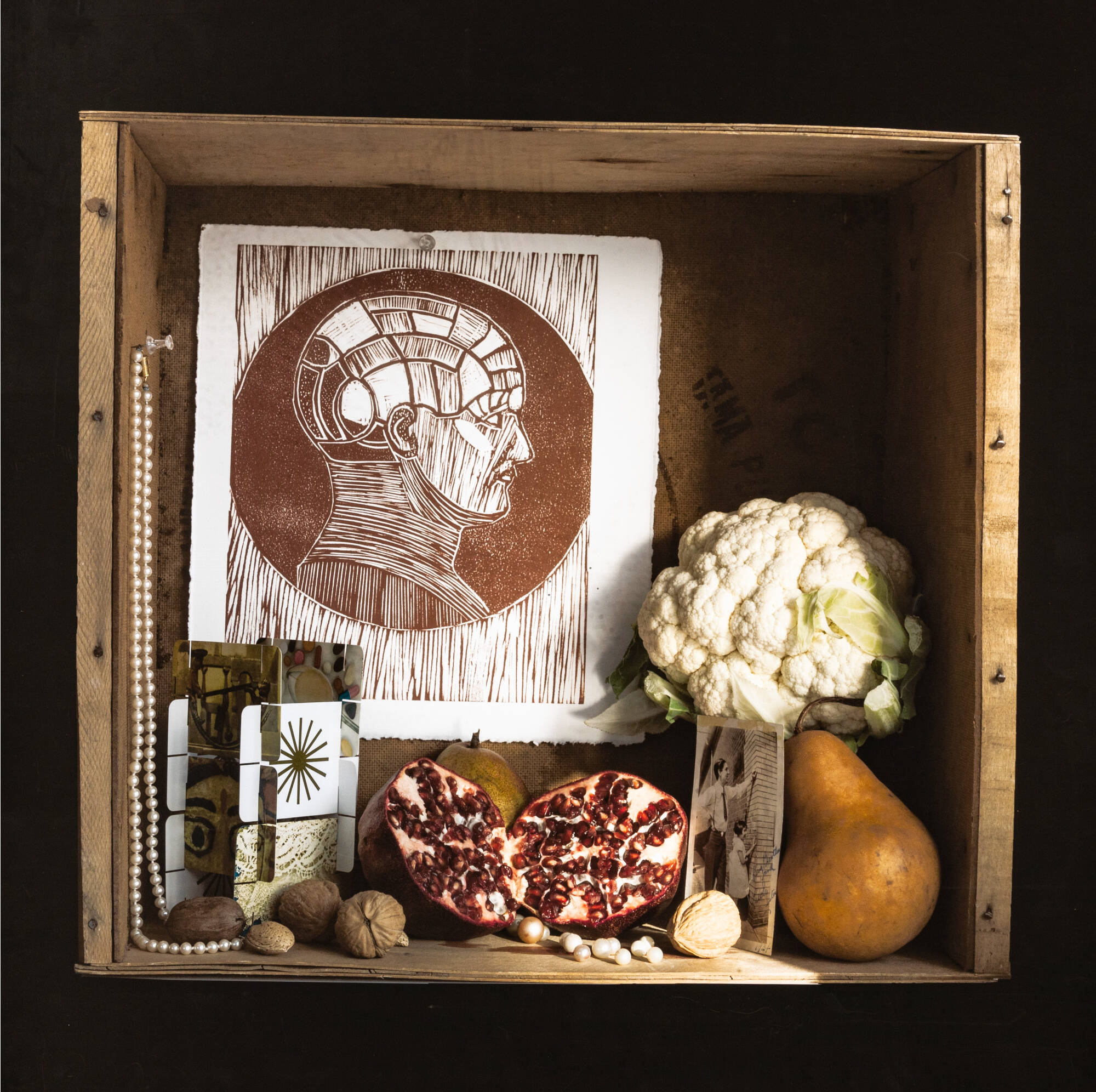 A shadowbox with a picture of a head in it along with a head of cauliflower, a cut in half pomegranate, a pear, and other trinkets.